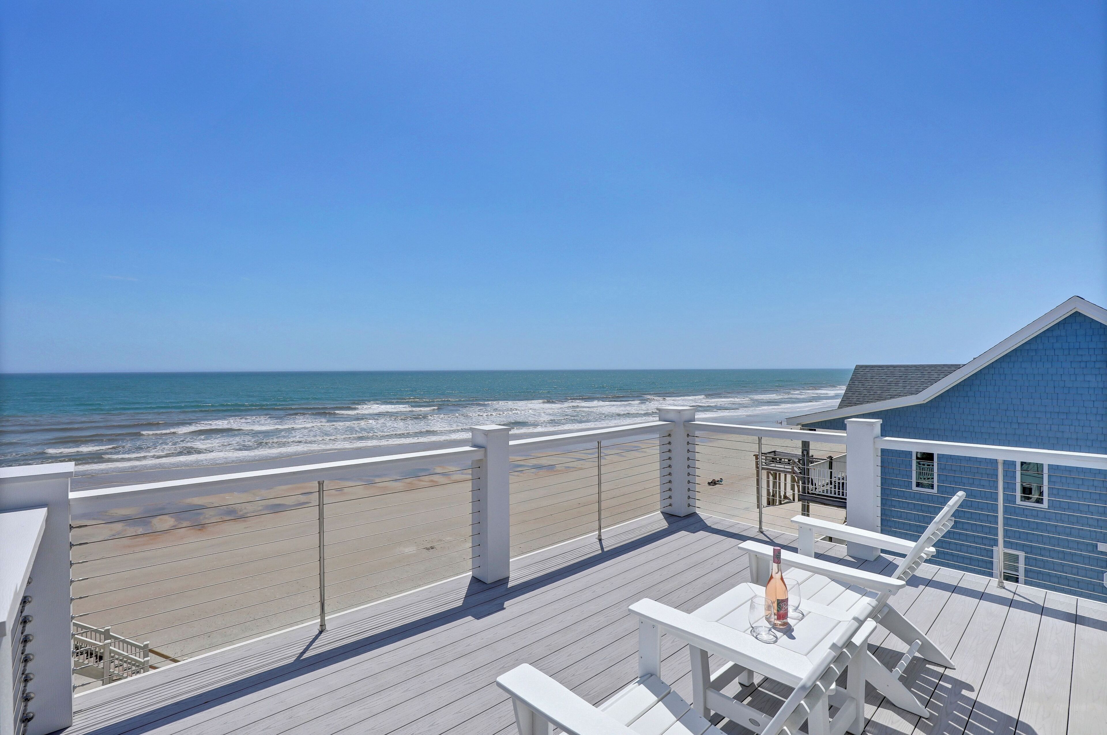 A beach view from a balcony with a wooden deck, a blue building, and a white railing.