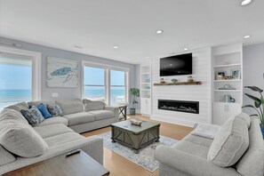 A modern living room with a large window overlooking the ocean, a fireplace, and a flat-screen TV mounted on the wall. - On Porpoise #223 | Luxury Oceanfront Escape in North Topsail Beach (North Topsail Beach)