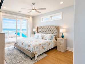 A bedroom with a large bed, a ceiling fan, a view of the beach, and a window with curtains. - On Porpoise #223 | Luxury Oceanfront Escape in North Topsail Beach (North Topsail Beach)