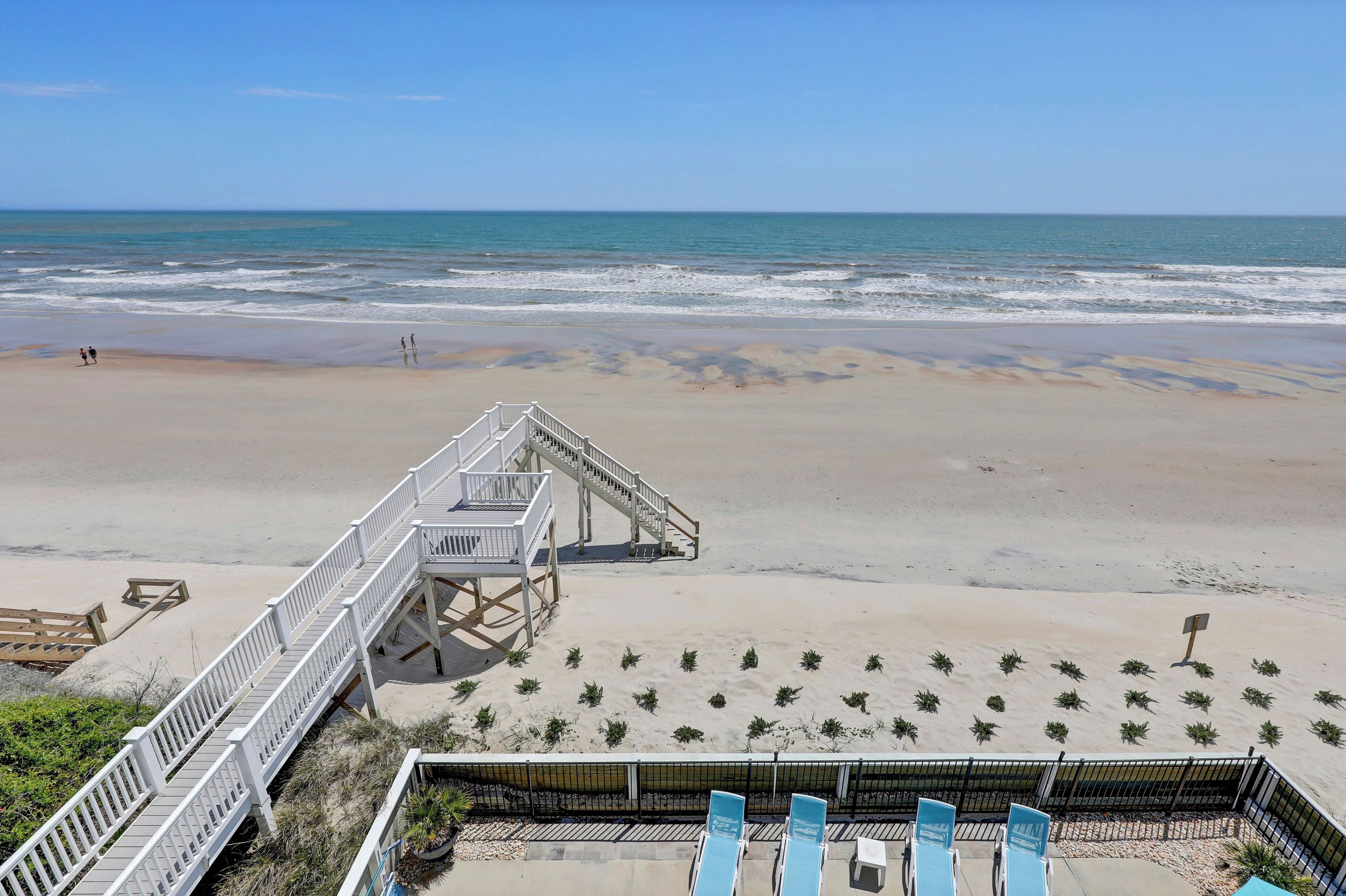 Une plage avec une promenade en bois menant à l’eau, une rangée de chaises bleues et un petit espace aménagé en jardin.