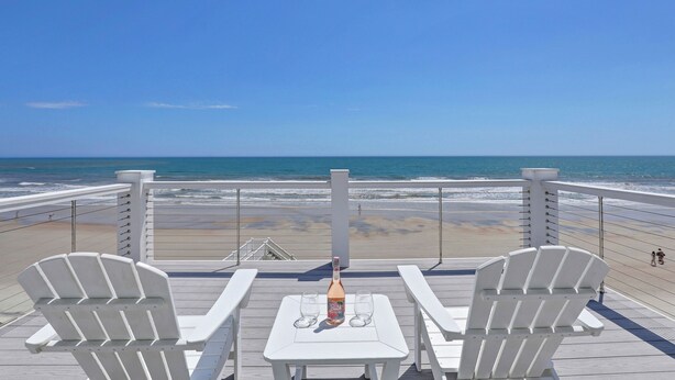 Eine Terrasse mit zwei weißen Stühlen und einem Tisch, mit Blick auf einen Strand und das dahinterliegende Meer.