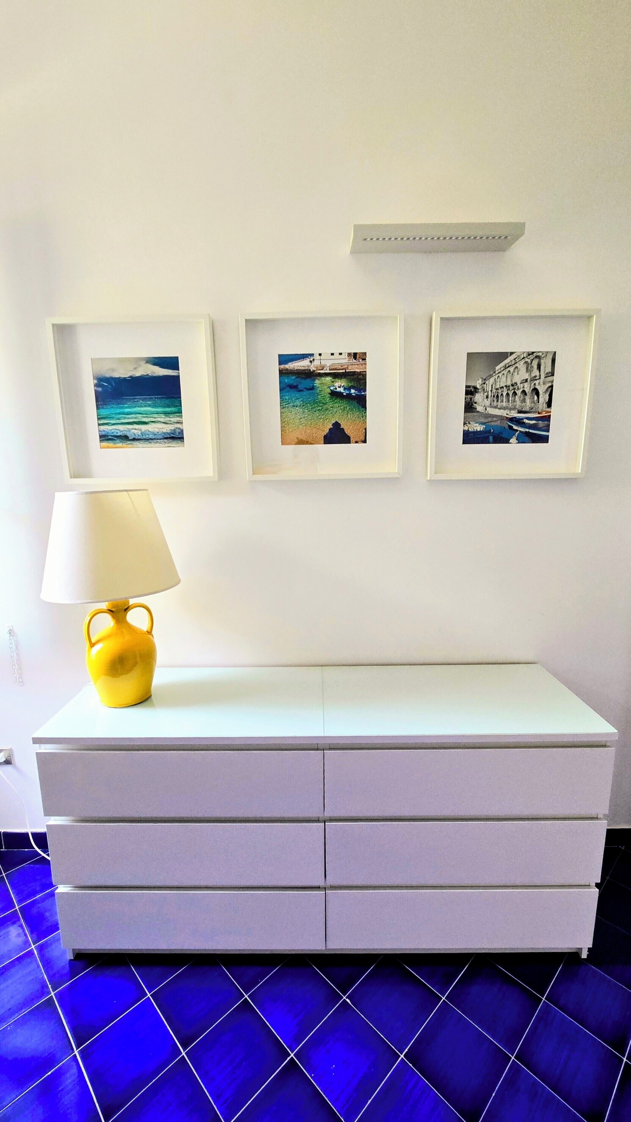 A white dresser with drawers, a yellow lamp, and three framed pictures on the wall.