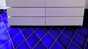 Valkoinen lipasto, jossa on laatikoita, keltainen lamppu ja kolme kehystettyä kuvaa seinällä.