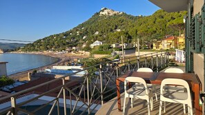 Ein Balkon mit Tisch und Stühlen mit Blick auf einen Strand und einen Hügel mit Gebäuden.