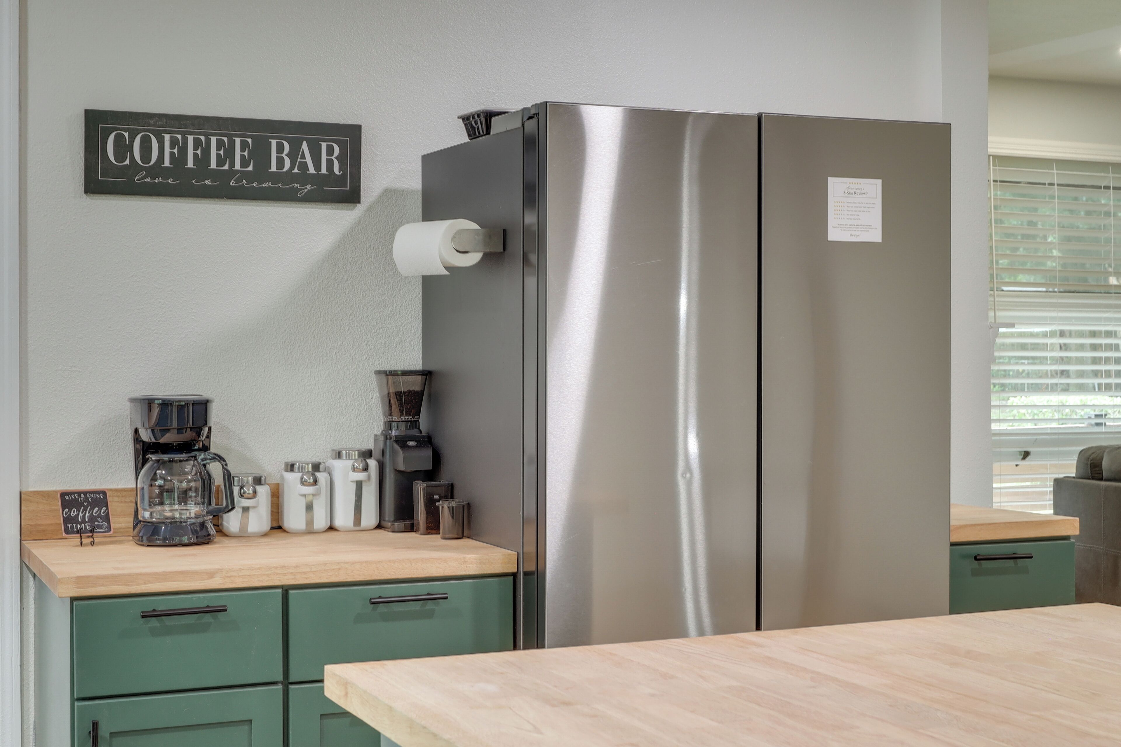Uma cozinha com geladeira de aço inoxidável, cafeteira e um letreiro que diz 'Coffee Bar love is brewing'.