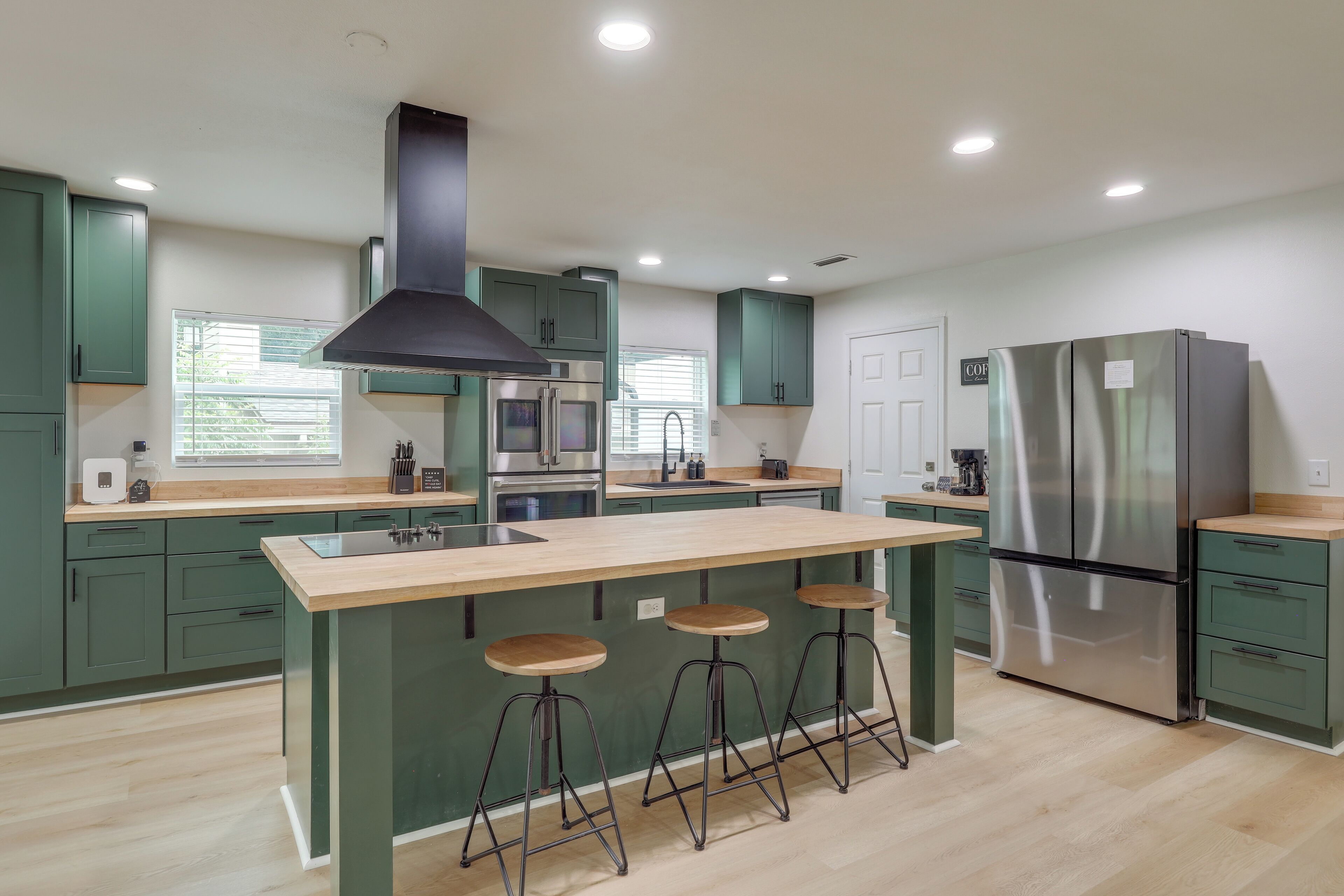 A modern kitchen with green cabinets, a stainless steel refrigerator, a central island with stools, and a black range hood.