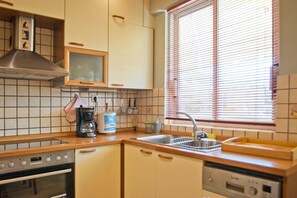A kitchen with a stove, oven, coffee maker, and a window with blinds. - Ellinikon Sea View (Argiroupoli)