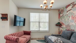 Una sala de estar con un sofá rosa, un sofá gris, un televisor de pantalla plana, una ventana con persianas y una pared con un mural floral.
