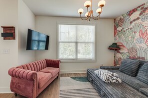 Ein Wohnzimmer mit einem rosafarbenen Sofa, einem grauen Sofa, einem Flachbildfernseher, einem Fenster mit Jalousien und einer Wand mit floralem Wandbild.