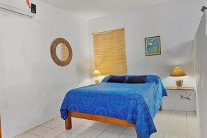 A bedroom with a bed, a round mirror, a window with blinds, a wall-mounted lamp, and a framed picture. - Quaint Condo Close to Beach and Plaza (Sayulita)