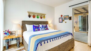 A bedroom with a bed, bedside tables, a lamp, a wall-mounted shelf with decorative items, and a door leading to an outdoor area. - Paraiso del Mar Casita: Walk to Beach & Pool! (La Paz)