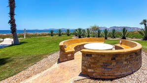 A circular stone fire pit with a seating area, surrounded by a well-maintained lawn and a view of the ocean and mountains in the background. - Paraiso del Mar Casita: Walk to Beach & Pool! (La Paz)
