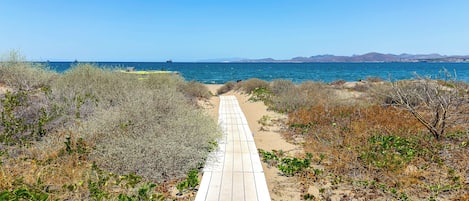 A wooden boardwalk leading to a beach with clear blue water and distant mountains.