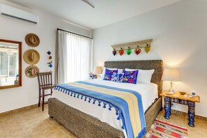 A bedroom with a bed, a chair, a bedside table, a lamp, a wall-mounted shelf with decorative items, and a window with curtains. - Paraiso del Mar Casita: Walk to Beach & Pool! (La Paz)