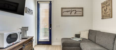 A compact living space with a kitchenette, a grey sofa, a wooden cabinet, and a door with blinds.