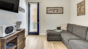 A compact living space with a kitchenette, a grey sofa, a wooden cabinet, and a door with blinds. - Steps to Empire Polo Club: Casita w/ Pool Access (Indio)