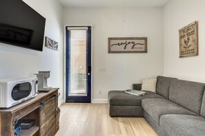 A compact living space with a kitchenette, a grey sofa, a wooden cabinet, and a door with blinds. - Steps to Empire Polo Club: Casita w/ Pool Access (Indio)