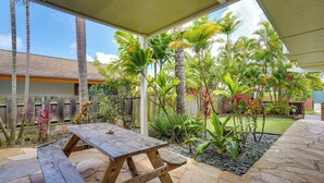Eira coberta com mesa e bancos de madeira, rodeada por plantas tropicais e um caminho de pedra.