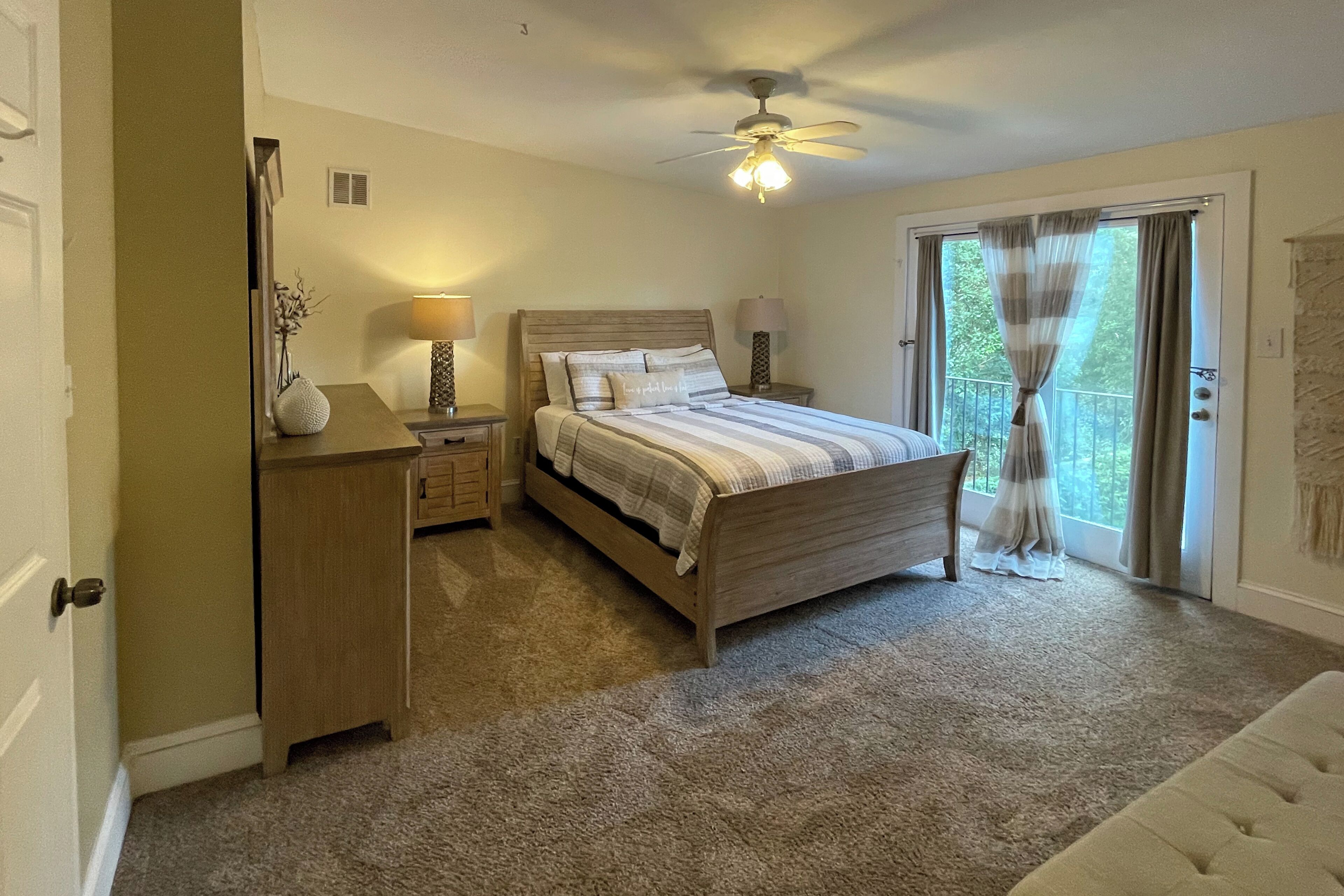 Un dormitorio con cama, lámparas de noche, mesita de noche, cómoda, ventilador de techo y puerta corredera de vidrio al exterior.