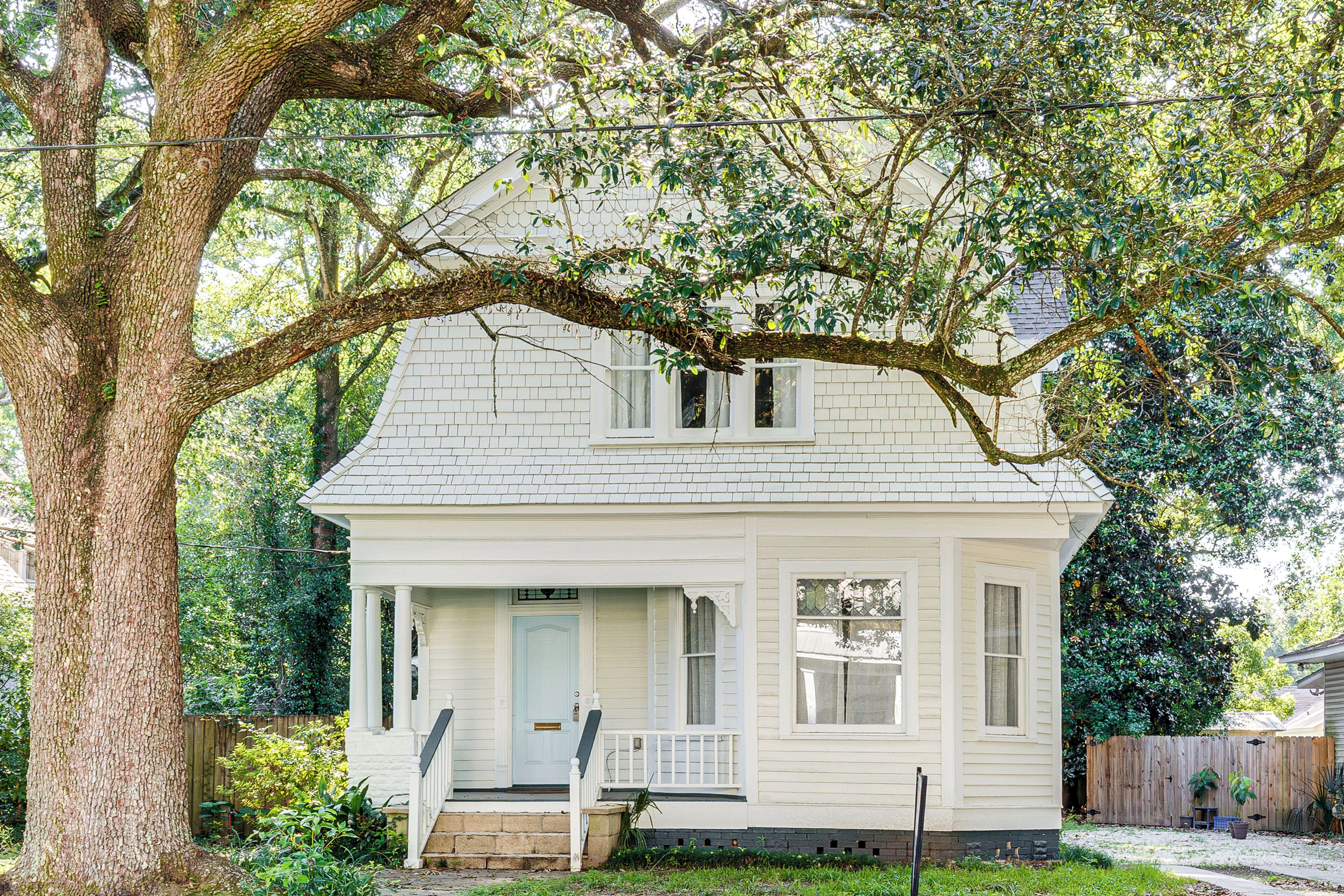 A white house with a porch, a front door, and a large tree in the background.