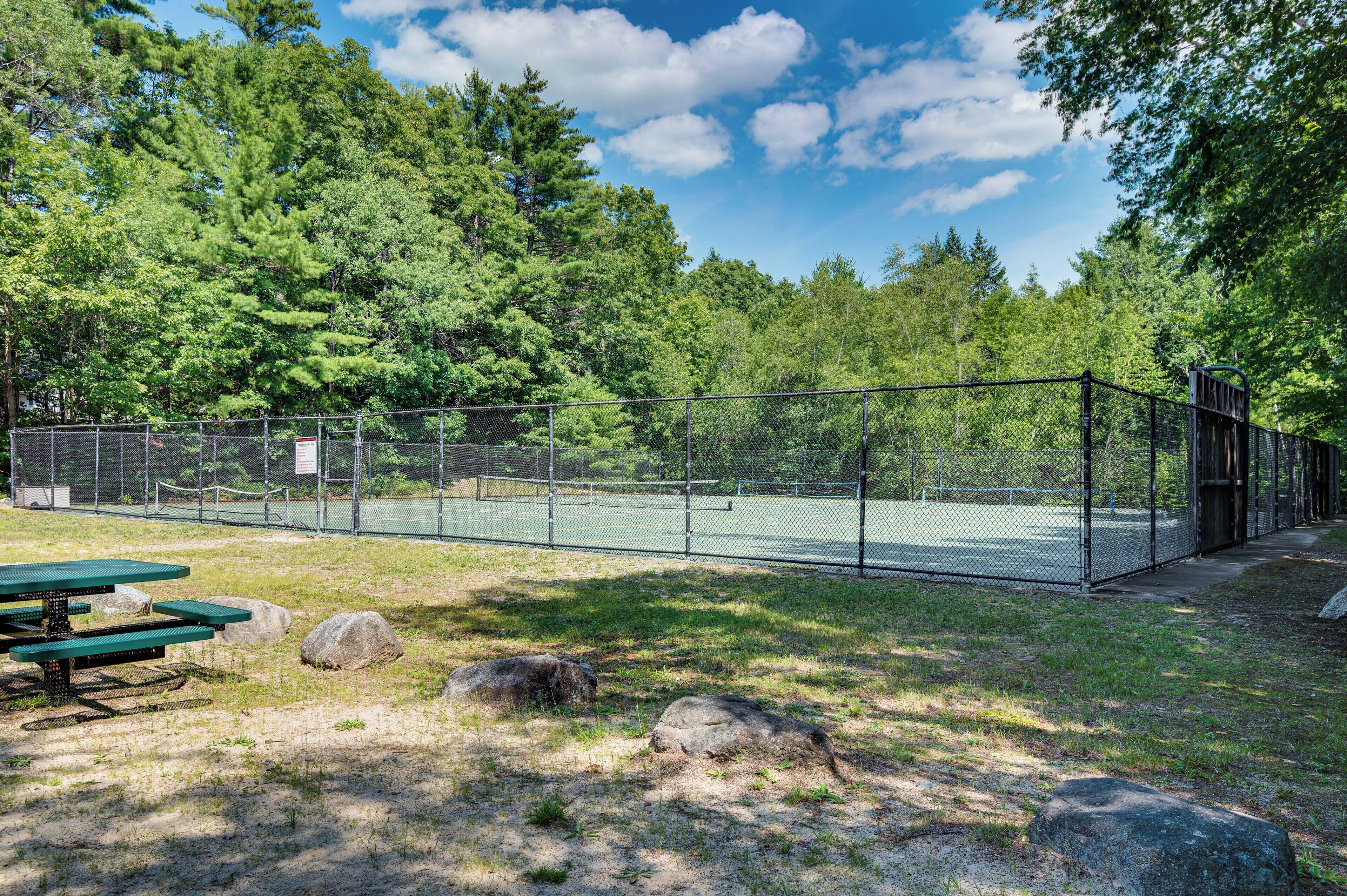 Un campo da tennis recintato con tavoli da picnic e rocce in una zona boschiva.