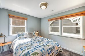 Ein Schlafzimmer mit einem Bett, zwei Fenstern mit Jalousien, einem Schreibtisch mit Lampe und Teppichboden.