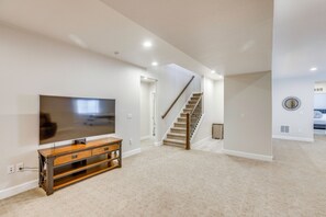 A spacious living room with a flat-screen TV on a wooden stand, a staircase leading to an upper level, and a kitchen area visible through an open doorway. - Great for Families! Arvada Haven w/ Yard & Patio (Arvada)