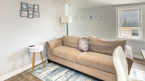 A living room with a beige sofa, a small side table, a lamp, and a window with blinds.
