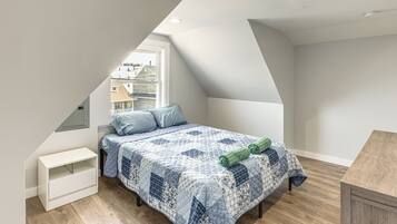 A bedroom with a sloped ceiling, a bed with a quilt, a nightstand, and a window with a view of houses.