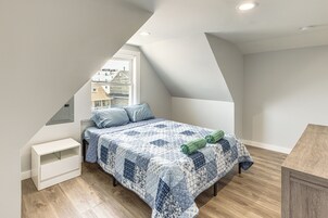 Un dormitorio con techo inclinado, una cama con colcha, una mesita de noche y una ventana con vista a casas.