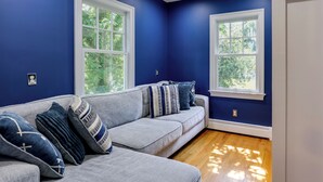 Una habitación con un sofá gris, dos ventanas y vista a árboles.