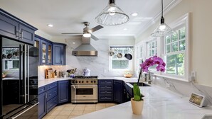 Una cocina moderna con armarios azul oscuro, encimera de mármol blanco, estufa de acero inoxidable y ventilador de techo.