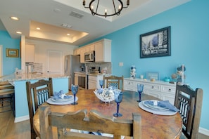 A dining area with a wooden table set for four, surrounded by chairs, in a room with blue walls and a kitchen in the background. - Waterfront Unit w/ Resort Perks in Laguna Vista! (Port Isabel)