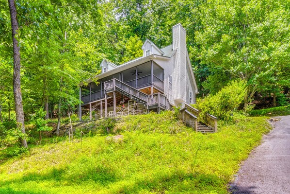 A two-story house with a balcony, surrounded by lush greenery and a forested area.