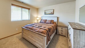 A bedroom with a wooden bed, a nightstand, a dresser, a window with blinds, and a painting on the wall. - Walk to Town! Charming La Conner Home (La Conner)