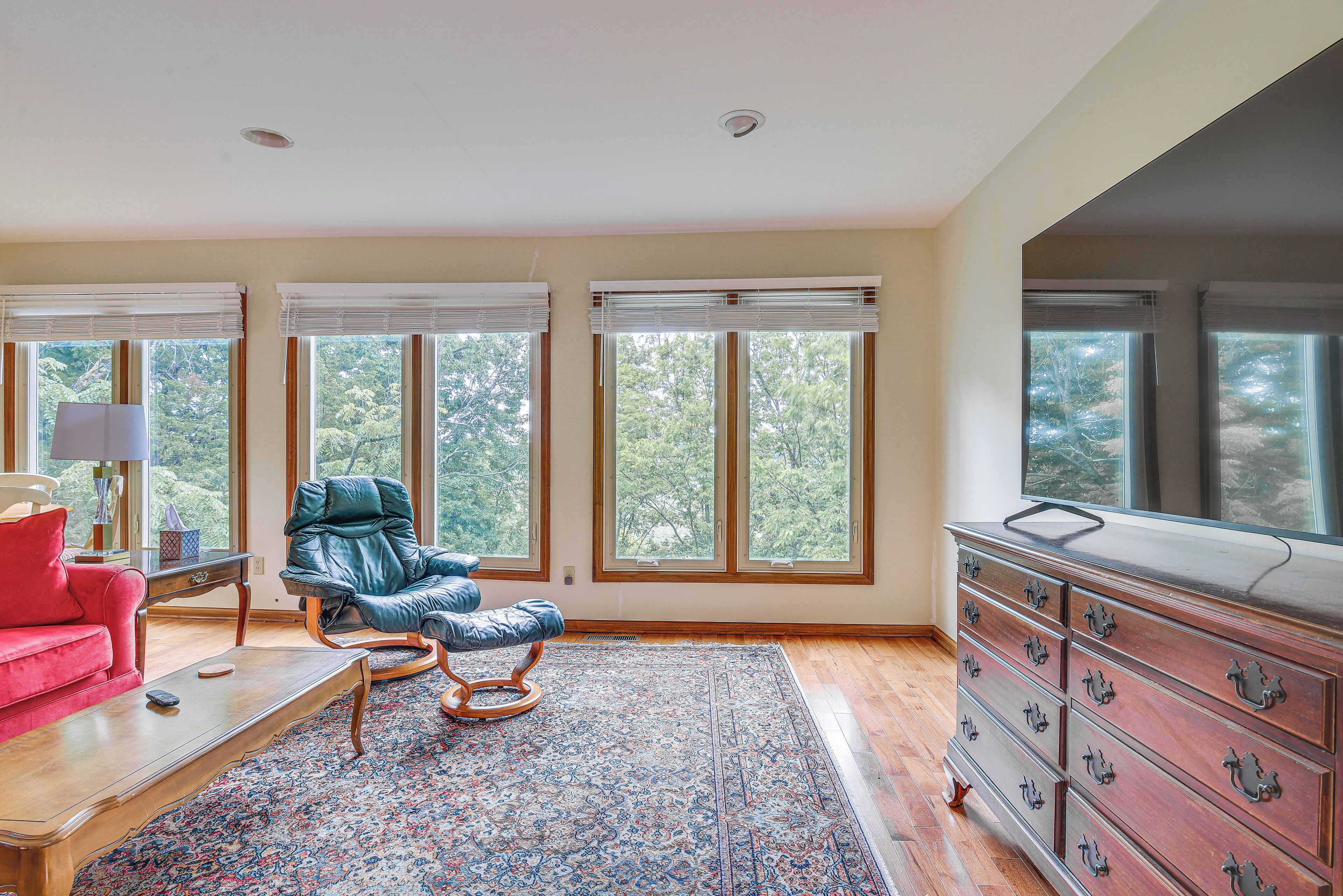 Une pièce avec un canapé rouge, une chaise bleue, une table basse en bois, un tapis à motifs, une commode avec de nombreux tiroirs et de grandes fenêtres équipées de stores.