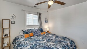 A bedroom with a bed, a ceiling fan, a window with curtains, a bedside table with a lamp, and a wall-mounted shelf with a framed picture and a sign.