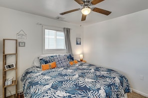 Ein Schlafzimmer mit Bett, Deckenventilator, Fenster mit Vorhängen, Nachttisch mit Lampe und Wandregal mit gerahmtem Bild und Schild.