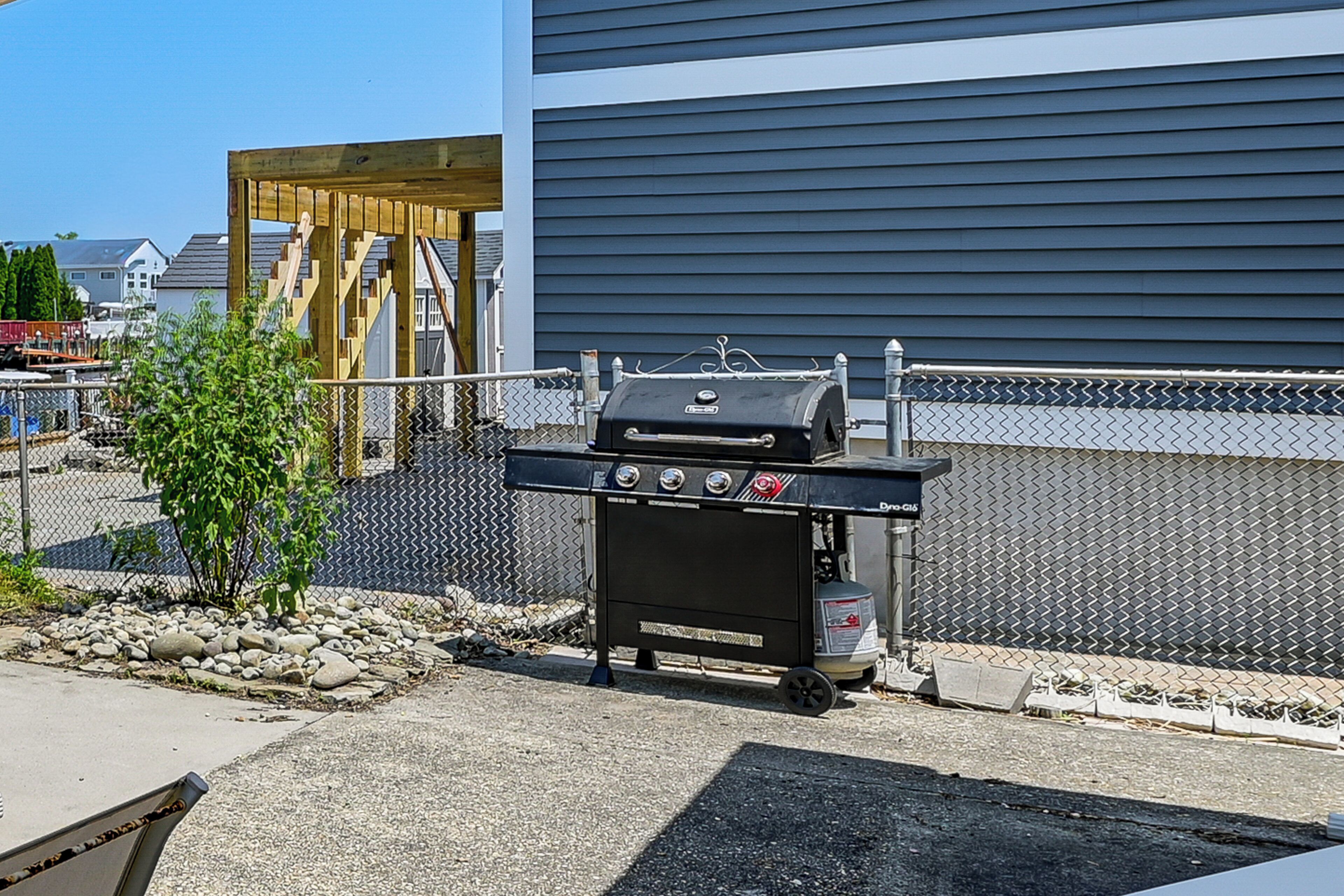 A barbecue grill with a gas tank and control knobs, positioned on a paved area with a chain-link fence and a wooden pergola in the background.