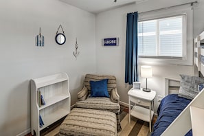 Un dormitorio con una cama nido, una cama individual, una mesita de noche con lámpara, una silla con un cojín azul, una estantería y adornos en la pared, incluyendo un espejo y un letrero.
