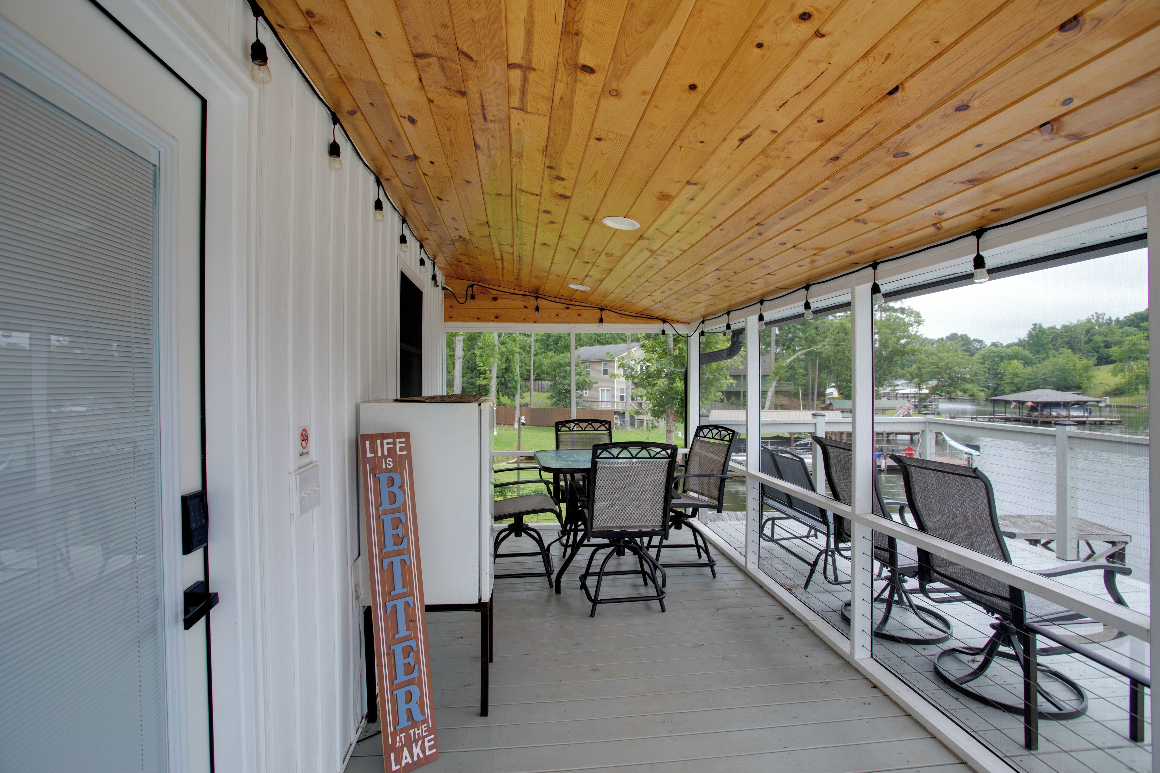 A covered outdoor seating area with a wooden ceiling, a refrigerator, and a sign that reads 'Life Better at the Lake.'