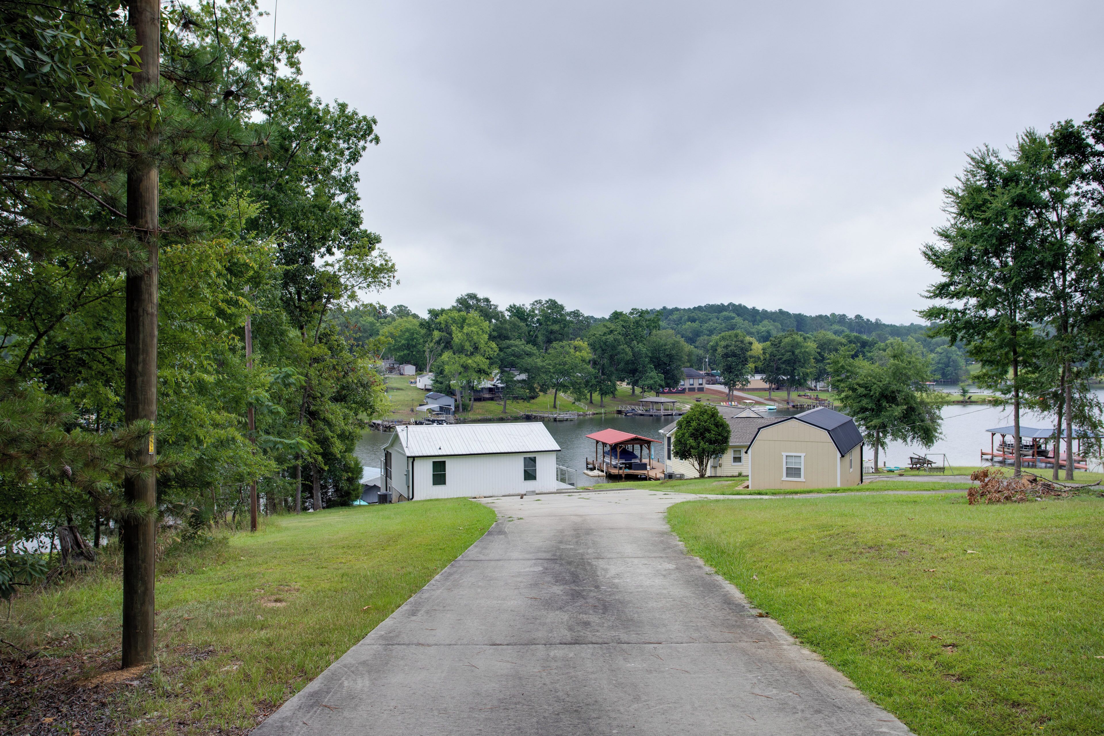 A pathway leading to a small building with a red roof, surrounded by greenery and a body of water in the background.