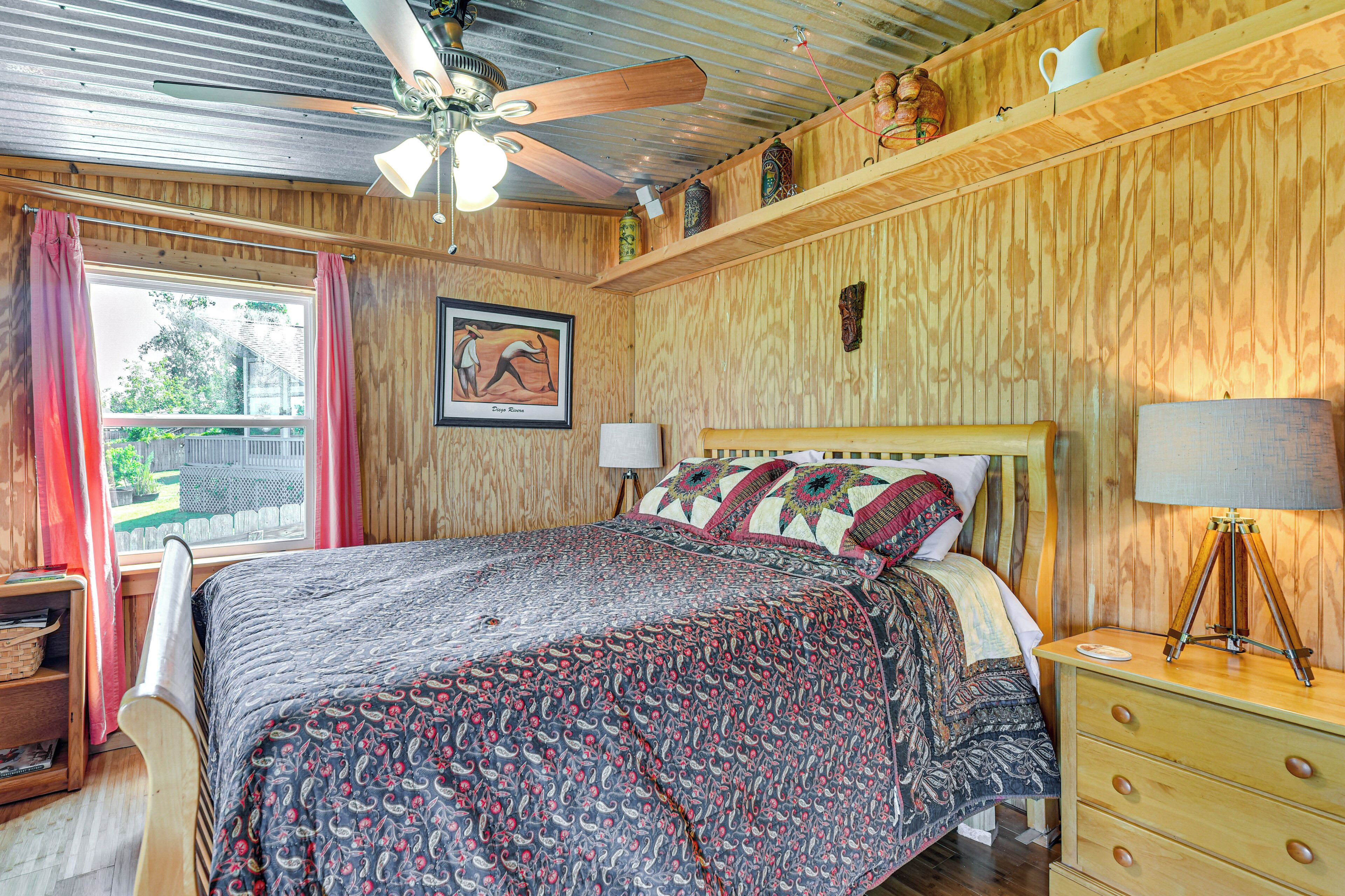 A bedroom with a bed, a ceiling fan, a window with curtains, a framed picture, and a bedside lamp.