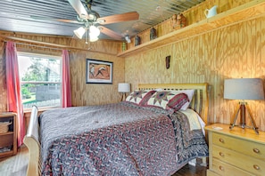 A bedroom with a bed, a ceiling fan, a window with curtains, a framed picture, and a bedside lamp.