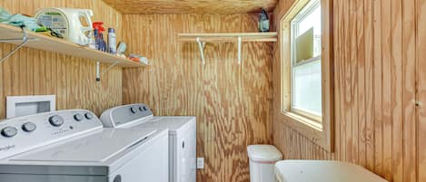 Un lavadero con lavadora y secadora de carga frontal, paredes de madera, una ventana y estanterías con productos de limpieza.