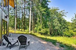A gravel patio with two Adirondack chairs, a wooden deck, and a forested backdrop. - Modern PNW Cabin on 10 Wooded Acres (Warren)