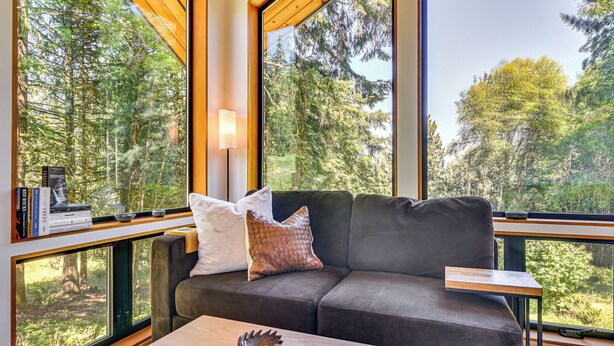 A modern living room with a grey sofa, a wooden coffee table, and a view of a forest through large windows. - Modern PNW Cabin on 10 Wooded Acres (Warren)