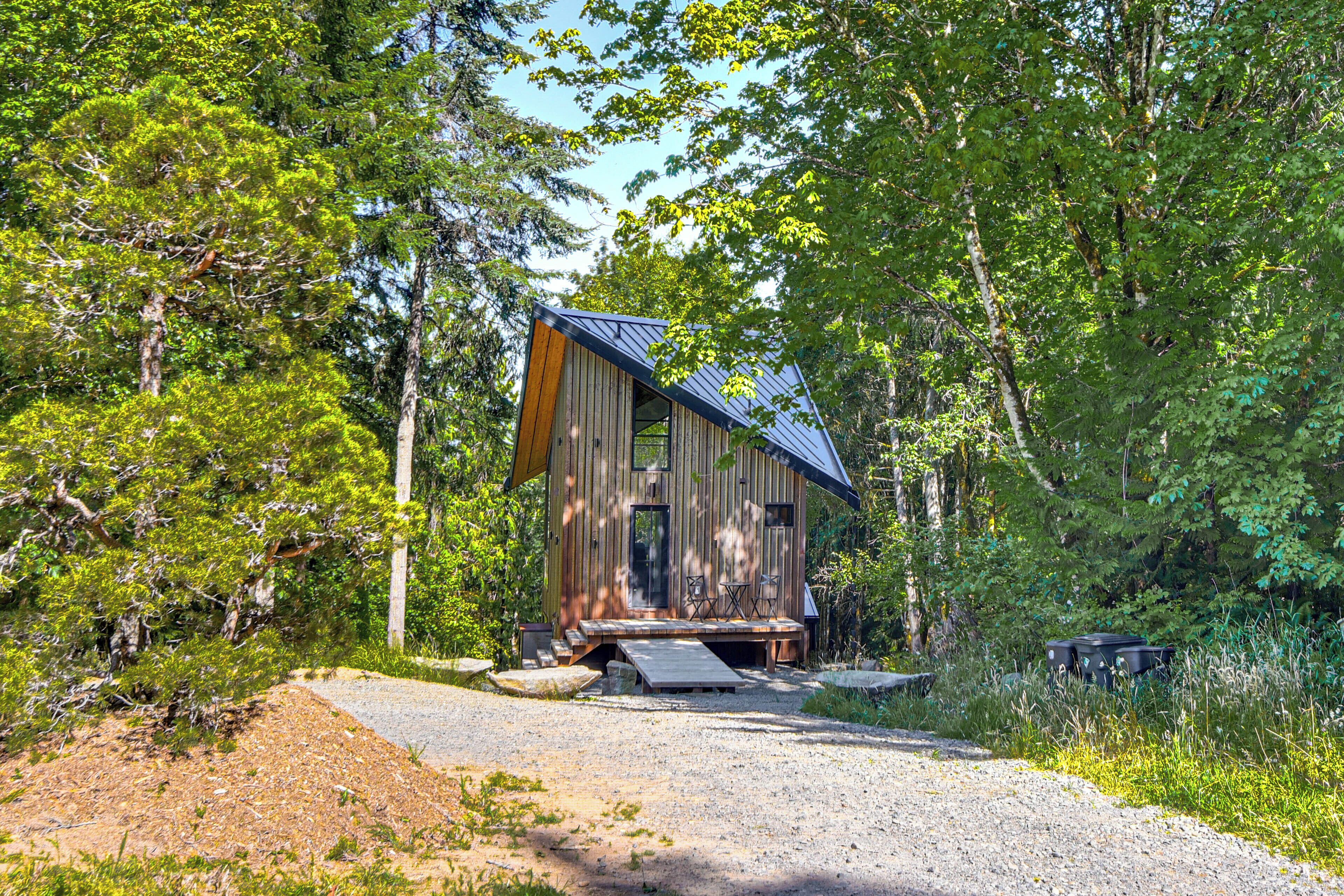 Un chalet rustique doté d’un toit en pente, d’une véranda avec des sièges et d’une allée en gravier entourée d’une forêt dense.