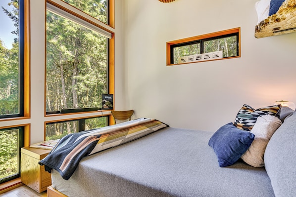 A bedroom with large windows, a bed with pillows, a bedside table, and a framed picture on the wall. - Modern PNW Cabin on 10 Wooded Acres (Warren)
