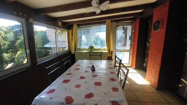 A dining area with a table covered in a floral tablecloth, wooden beams on the ceiling, a window with a view of greenery, and a clock on the wall. - The Symphony of the Birds (Villelongue-d'Aude)